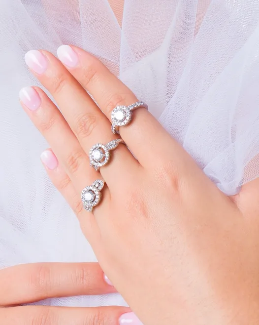 Trois bagues à pierre ronde entourée d'un halo de petites pierres, sur une main posée sur un tulle blanc